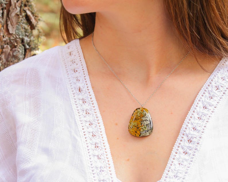 Model wearing a botanical resin necklace with real lichens in a warm-toned pendant. The piece stands out against a white embroidered top and natural outdoor background, showcasing its wearable scale