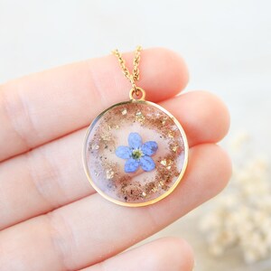 Puede incluir: Un collar dorado con un colgante redondo. El colgante presenta una peque&ntilde;a flor azul encapsulada en resina transparente, rodeada de copos dorados. El collar est&aacute; sostenido en una mano.