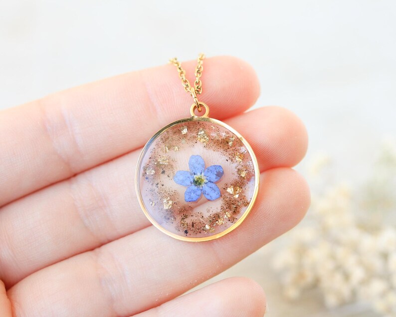 Puede incluir: Un collar dorado con un colgante redondo. El colgante presenta una peque&ntilde;a flor azul encapsulada en resina transparente, rodeada de copos dorados. El collar est&aacute; sostenido en una mano.