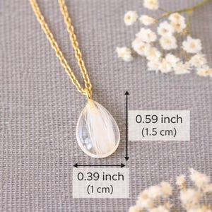 Pode incluir: Colar dourado com pingente em forma de gota. O pingente transparente cont&eacute;m detalhes brancos e mede 1,5 cm de altura e 1 cm de largura. O colar &eacute; exibido em um fundo cinza.