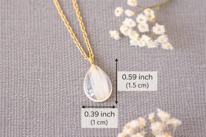 Pode incluir: Colar dourado com pingente em forma de gota. O pingente transparente cont&eacute;m detalhes brancos e mede 1,5 cm de altura e 1 cm de largura. O colar &eacute; exibido em um fundo cinza.
