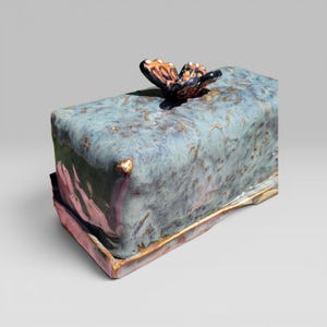 May include: A rectangular ceramic butter dish with a teal glaze and gold accents. A detailed butterfly with orange, black, and white markings sits atop the lid. The dish has a pink and gold base.