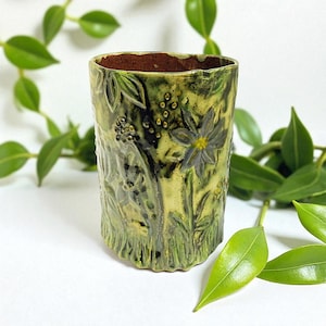 May include: A handmade ceramic vase with a textured finish and a botanical design. The vase features embossed flowers, leaves, and stems in shades of green, yellow, and brown. The interior is a dark brown colour.