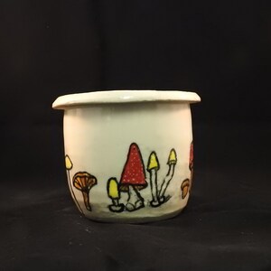 May include: A small, off-white ceramic pot with a rounded shape and a slightly flared rim. The pot is decorated with hand-painted mushrooms in red, yellow, and brown, with black outlines. The mushrooms are arranged around the base of the pot.