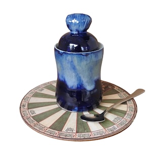 May include: A blue and white ceramic jar with a lid, sitting on a round wooden tray with a decorative design. A silver spoon is on the tray.