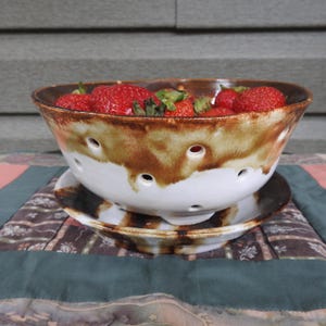 May include: A ceramic berry bowl and saucer, filled with fresh red strawberries. The bowl is white with brown glaze accents and drainage holes. The bowl and saucer sit on patterned fabric.