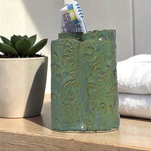May include: A green ceramic toothbrush holder with an embossed floral design. A tube of toothpaste and a toothbrush are placed inside the holder. The holder is on a wooden surface, with a succulent plant and white towels in the background.