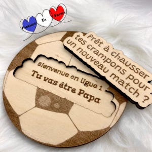Può includere: Targa in legno a forma di pallone da calcio con un ritaglio per un messaggio. La targa presenta il testo "Bienvenue en ligue 1 Tu vas être Papa" e "Prêt à chausser tes crampons pour un nouveau match ?" in marrone scuro. Sono presenti anche le parole "Fabriqué en France".