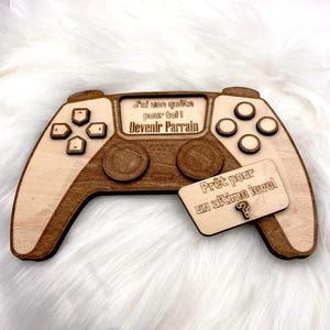 Può includere: Targa in legno a forma di controller di gioco con testo inciso. La targa è marrone chiaro con accenti marrone scuro. Presenta pulsanti, pad direzionali e due pad di controllo circolari. Un'etichetta più piccola recita "Prêt pour un ultime level".