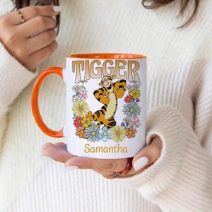 May include: White ceramic mug with an orange rim. The mug features a cartoon image of Tigger from Winnie the Pooh, surrounded by colourful flowers. The text "Tigger" is printed in orange above the image, and the name "Samantha" is printed below the image.