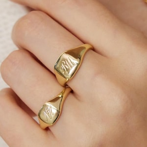 Op de afbeelding: Twee gouden zegelringen met gegraveerde initialen worden aan een hand gedragen, naast een fijne gouden ketting. De ringen hebben een gepolijste afwerking en een klassiek, elegant ontwerp. De achtergrond is zacht off-white.