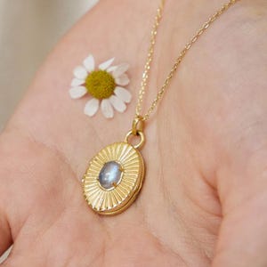 May include: A gold-colored necklace with a pendant featuring a blue-grey stone is displayed on an open hand. The pendant is oval-shaped with a sunburst design. A small white daisy with a yellow center rests near the necklace.