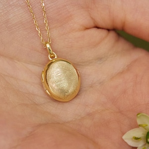 Könnte beinhalten: Eine goldfarbene ovale Medaillon-Halskette mit strukturierter Oberfläche, die an einer feinen Kette hängt. Eine kleine weiß-grüne Blume liegt in einer Hand neben der Halskette.