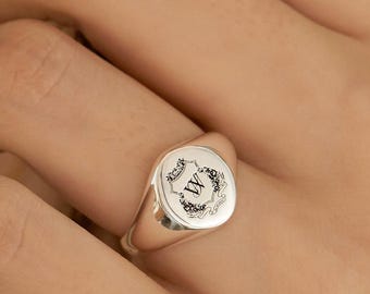 Peut inclure: Bague chevalière en argent avec une face circulaire ornée d'une couronne florale et de la lettre "W". La bague est portée au doigt d'une personne, avec une vue rapprochée mettant en évidence les détails du design.