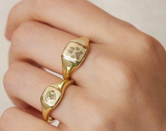 May include: Two gold signet rings are worn on a hand. One ring features a paw print design, and the other has a small, circular design. A gold necklace with a small pendant is also visible.