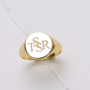 May include: A gold signet ring featuring a flat, circular face engraved with the letters T, S, and R in a stacked, serif font. The ring has a polished, reflective metallic finish and a smooth, tapered band that connects to the circular top.