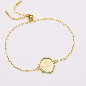 Op de afbeelding: Een goudkleurige verstelbare armband met een fijne ketting en een centrale, onregelmatig gevormde ronde bedel. De bedel is gegraveerd met de cursieve letters LR. De armband heeft een schuifkraalmechanisme om de lengte aan te passen.