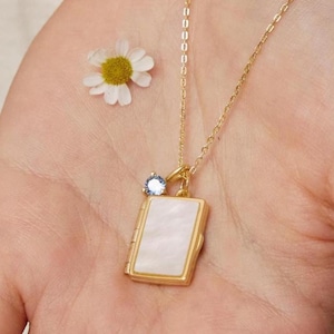 Könnte beinhalten: Eine goldfarbene Halskette mit einem rechteckigen Anhänger mit weißer, schillernder Oberfläche. Ein kleiner, hellblauer Edelstein und eine Gänseblümchen sind ebenfalls zu sehen. Die Halskette wird in einer offenen Hand gehalten.