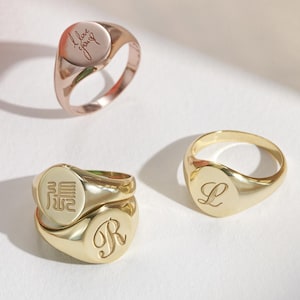 May include: Three gold signet rings with engraved designs and a rose gold ring with a script design are displayed on a white surface. The rings have different engravings, including a letter and script. A pomegranate and a vase are in the background.