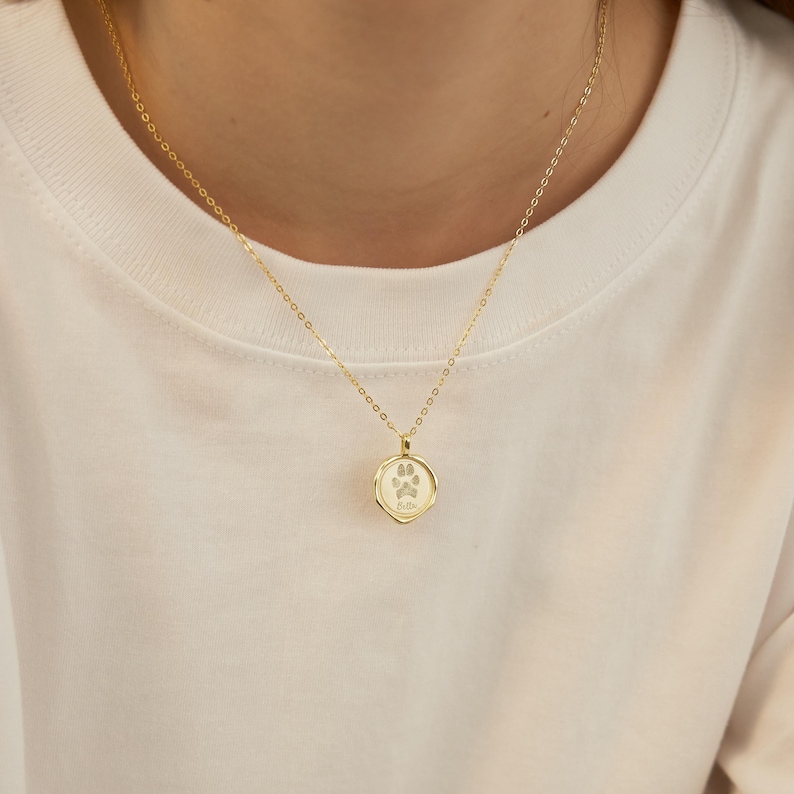 Puede incluir: Un collar de cadena de oro con un peque&ntilde;o colgante de oro con una huella de pata. El colgante es redondo y tiene una superficie ligeramente texturizada.