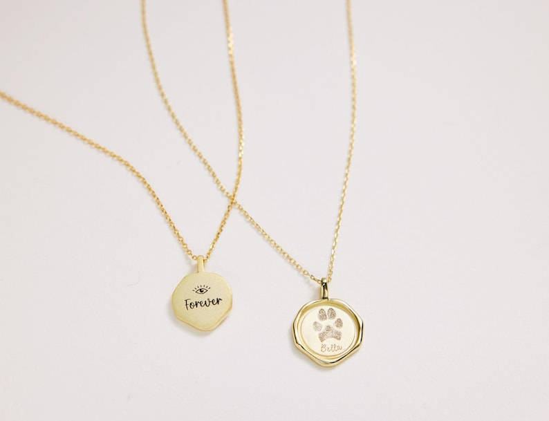Puede incluir: Dos collares de oro con colgantes. Un colgante tiene la palabra "Forever" y un s&iacute;mbolo de ojo. El otro colgante tiene una huella de pata y la palabra "Bella".