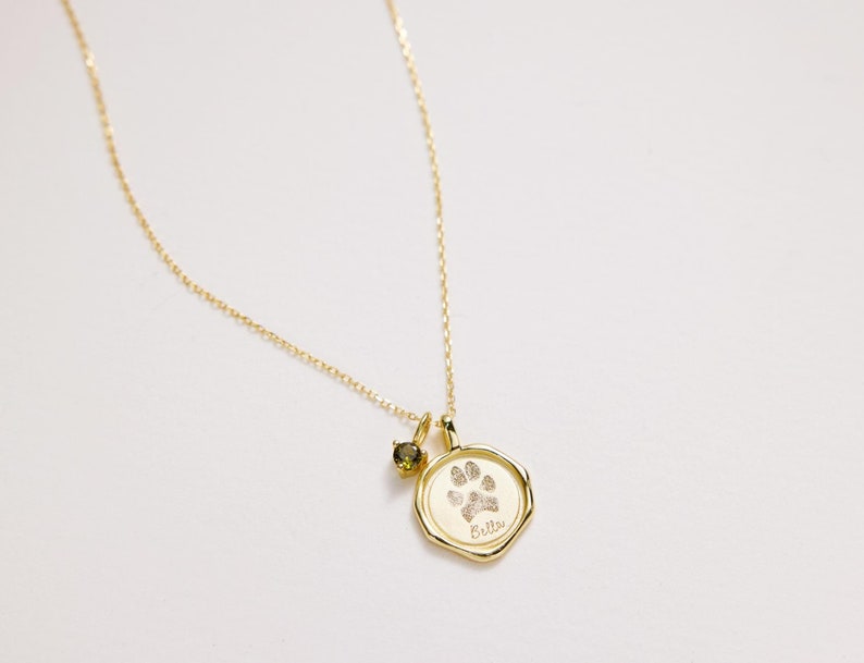 Puede incluir: Collar de oro con un dije de huella de pata y una peque&ntilde;a piedra verde. El dije tiene el nombre "Bella" grabado.