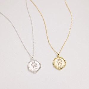 Puede incluir: Dos collares de plata y oro con dijes de huella de pata. El collar de plata tiene el nombre "Faye" grabado en el dije. El collar de oro tiene el nombre "Bella" grabado en el dije.