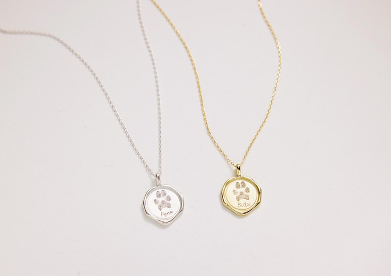 Puede incluir: Dos collares de plata y oro con dijes de huella de pata. El collar de plata tiene el nombre "Faye" grabado en el dije. El collar de oro tiene el nombre "Bella" grabado en el dije.