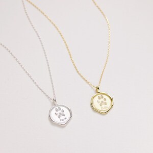 Puede incluir: Dos collares de plata con colgantes de huella de pata. El colgante del collar de la izquierda dice "Kyla" y el colgante del collar de la derecha dice "Bella".