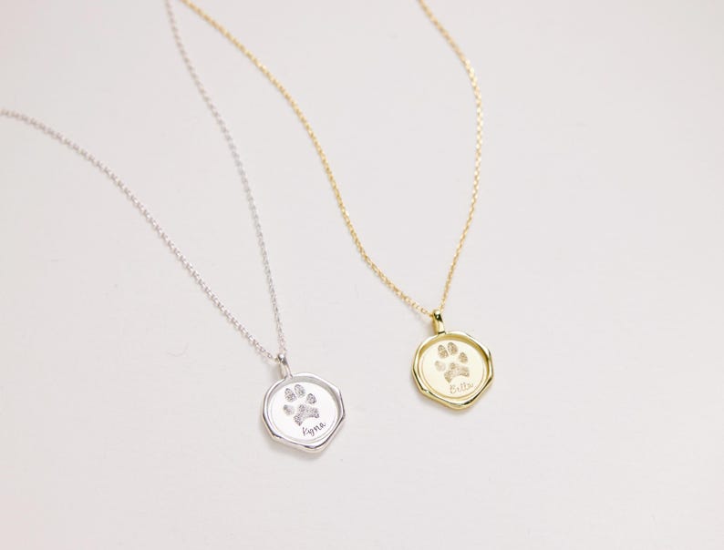 Puede incluir: Dos collares de plata con colgantes de huella de pata. El colgante del collar de la izquierda dice "Kyla" y el colgante del collar de la derecha dice "Bella".