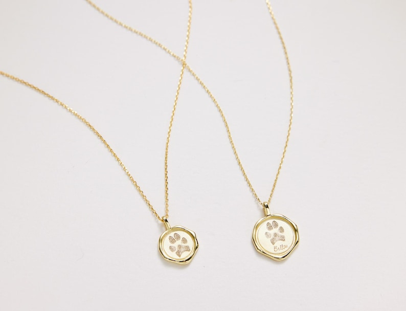 Puede incluir: Dos collares de oro con colgantes de huella de pata. Los colgantes son redondos y tienen un dise&ntilde;o de huella de pata en relieve. Un colgante tiene la palabra "Bella" grabada.