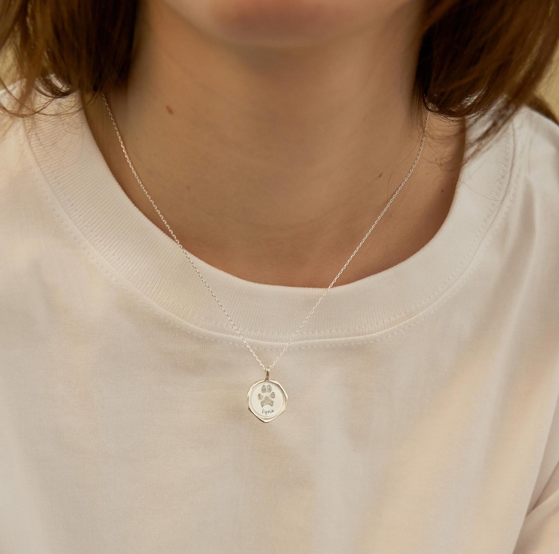 Puede incluir: Un collar de plata con un peque&ntilde;o colgante redondo que presenta una huella de pata. El colgante est&aacute; grabado con la palabra "Aspen".