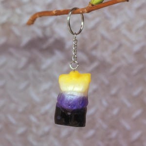 May include: A non-binary pride flag keychain. The charm, shaped like a torso, displays the flag's colours: yellow, white, purple, and black. It hangs from a silver chain and ring, suspended from a small branch, against a textured background.