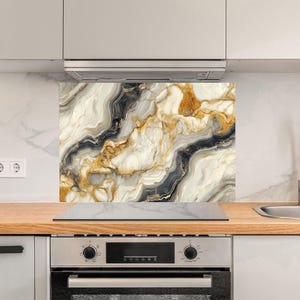 Könnte beinhalten: Eine Küchenrückwand mit Marmormuster in Weiß-, Schwarz- und Goldtönen. Die Rückwand befindet sich hinter einem schwarzen Kochfeld und Backofen.
