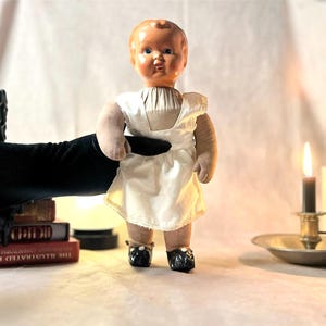 May include: A vintage doll with a painted face and a white dress. The doll is standing in front of a stack of books and a lit candle. The doll is wearing black shoes. A black gloved hand is holding the doll.
