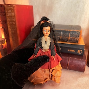 May include: A vintage doll in a traditional Spanish costume. The doll has black hair, a black veil, and a red and orange layered skirt. The doll is held in a black velvet glove, with antique books in the background.