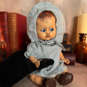 Puede incluir: Una muñeca bebé vintage con ojos azules, vestida con un vestido y gorro azul claro. La muñeca tiene zapatos marrones y está sujeta por una mano enguantada de negro. La muñeca está sentada.