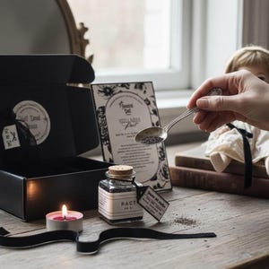 Puede incluir: Una caja negra abierta con una etiqueta que dice "Still Dead" revela una pequeña bolsa. Un pequeño frasco de vidrio etiquetado "PACT" se encuentra junto a una vela rosa encendida. Una muñeca y una cuchara con semillas también están en la imagen.