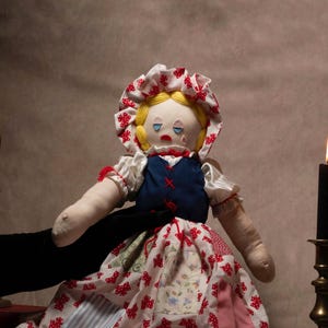 May include: A handmade rag doll with a bonnet and patchwork dress. The doll has blonde yarn hair, blue eyes, and a red teardrop. The dress is white with red polka dots and various fabric patches. The doll is held by a black-sleeved arm.