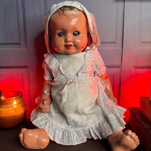 May include: A vintage composition doll with a glossy peach-toned face, blue eyes, and painted red lips. The doll wears a white bonnet and a matching short-sleeved dress featuring a subtle floral pattern, ruffled trim, and a tie at the waist.