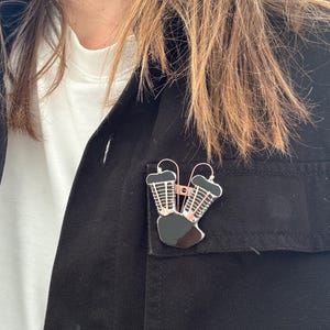 Può includere: Uno spillo a forma di motore nero e argento con finitura oro rosa. Lo spillo è indossato su una giacca nera.