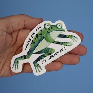 Könnte beinhalten: Ein Aufkleber mit einem grünen Frosch mit schwarzen Flecken und Streifen. Der Frosch befindet sich auf einem weißen Hintergrund mit dem deutschen Text "SPRING EIN FÜR DIE DEMOKRATIE" in Schwarz. Der Aufkleber wird in einer Hand vor blauem Hintergrund gehalten.