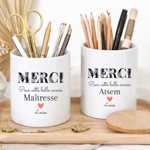 Può includere: Due portapenne in ceramica bianca con la scritta "MERCI" in caratteri floreali neri. Uno dice "Maîtresse" e l'altro "Atsem". Entrambi hanno un cuore rosso e il nome "Louise". I portapenne sono su un vassoio di legno con articoli di cancelleria.