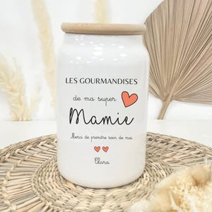 Peut inclure: Un pot en céramique blanc avec un couvercle en bois, portant l'inscription "LES GOURMANDISES de ma super Mamie" et un cœur rouge. Le pot comprend également le texte "Merci de prendre soin de moi" et le nom "Clara".