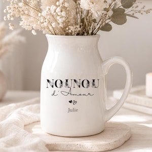 Peut inclure: Un pichet en céramique blanc avec une anse, rempli de fleurs séchées et de verdure. Le pichet porte l'inscription "NOUNOU d'Amour", le nom "Julie" et un symbole de cœur. Une bougie allumée est visible en arrière-plan.