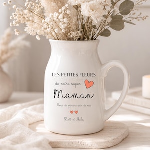Peut inclure: Un vase en céramique blanc avec une anse, rempli de fleurs séchées et de verdure. Le vase porte du texte en français, dont le mot "Maman" et un symbole de cœur. Une bougie allumée et un petit vase sont en arrière-plan.