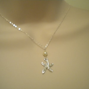 May include: A silver necklace featuring a starfish pendant. The pendant is detailed with small embellishments and hangs below a small, round, gold-colored bead. The necklace chain is delicate and silver.