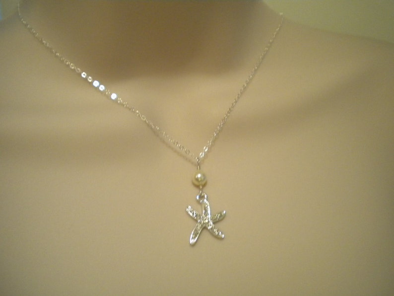 May include: A silver necklace featuring a starfish pendant. The pendant is detailed with small embellishments and hangs below a small, round, gold-colored bead. The necklace chain is delicate and silver.