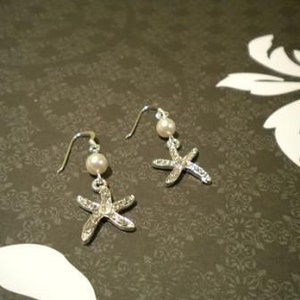May include: A pair of silver-toned starfish earrings with a pearl bead and a hook closure. The starfish are encrusted with small, clear crystals. The earrings are displayed on a dark background with a floral pattern.