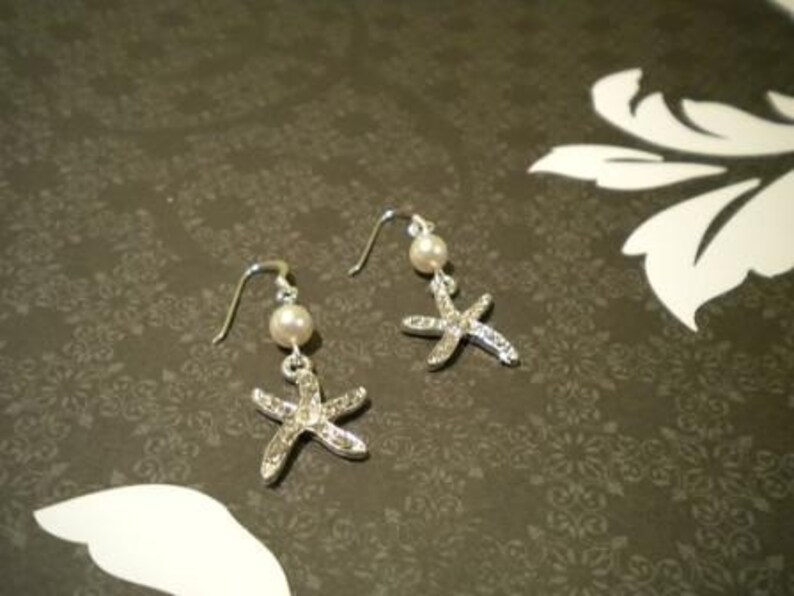 May include: A pair of silver-toned starfish earrings with a pearl bead and a hook closure. The starfish are encrusted with small, clear crystals. The earrings are displayed on a dark background with a floral pattern.